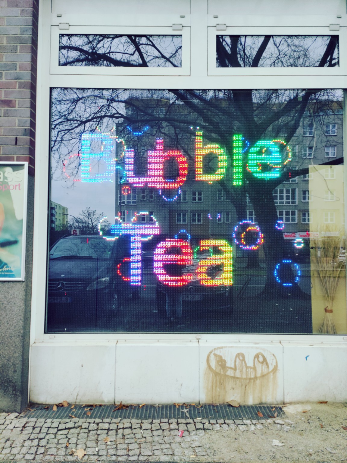 Schaufenster in einer Berliner Straßenszene mit Spiegelungen von Bäumen, Häusern und Autos.